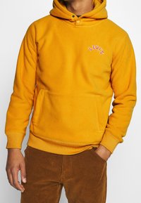 Sudadera con capucha de forro polar amarillo con un bolsillo frontal y el logo "LEVI'S" bordado en rosa. Diseñada con capucha ajustable con cordones y puños acanalados.