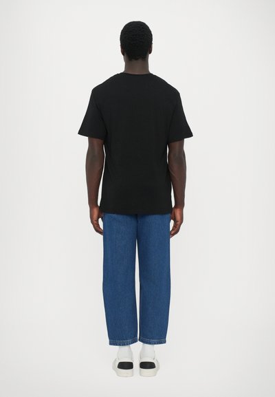 Black cotton t-shirt with short sleeves, seen from the back, paired with blue denim pants and white sneakers with black accents.