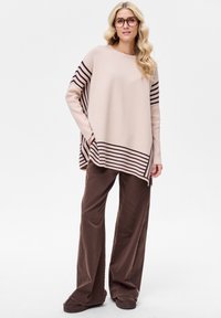 Lichtroze gebreide poncho met donkere bruine horizontale strepen, gecombineerd met losse bruine broek en donkere instapschoenen. Soepele textuur.