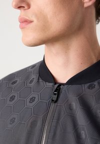Gray bomber jacket with a hexagonal pattern, featuring a black collar and zipper, constructed from smooth, lightweight fabric.