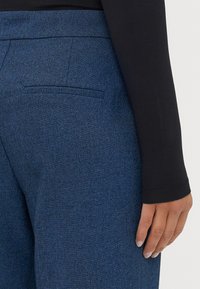 Donkerblauwe op maat gemaakte broek van een subtiele gestructureerdestof, met een achterzak en een close-up van een zwart lange mouwen top.