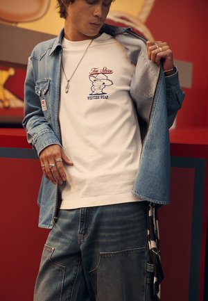 Jeansowa kurtka na białym t-shircie z rysunkiem i tekstem, w połączeniu z jasnoniebieskimi, luźnymi dżinsami. Dodatki to pierścionki i naszyjnik z zawieszką.