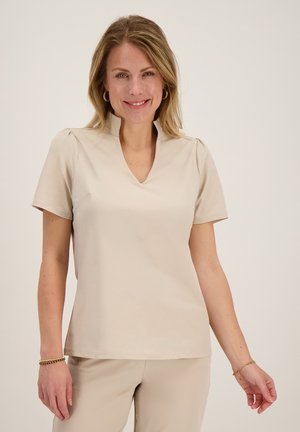 Glimlachende vrouw met blond haar, die een beige top met korte mouwen en bijpassende broek draagt, staand tegen een effen lichte achtergrond.