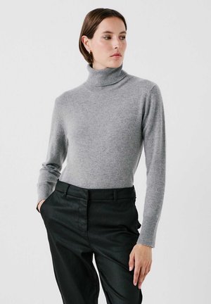 Pull à col roulé gris en tricot doux, doté de manches longues ajustées et d'une texture lisse. Associé à un pantalon noir près du corps.