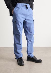 Calças cargo azuis com cintura elástica e cordão. Apresentam múltiplos bolsos laterais e um ajuste descontraído. Usadas com sapatos pretos de atar.