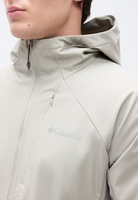 Gros plan d'une personne portant une veste zippée Columbia gris clair avec capuche et poche poitrine zippée.