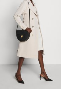 Trench beige con dettagli a bottoni, abbinato a una borsa a tracolla in pelle nera con chiusura dorata. Il look è completato da décolleté nere con tacco a spillo.