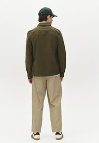 Jachetă de catifea verde măsliniu, pantaloni bej, vedere din spate. Caracteristici includ un guler cu nasturi, croială dreaptă și adidași clasici cu accente negre.