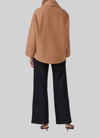 Unâ giacca over-size in misto lana color beige con colletto a punte, spalle scese e orlo curvo, abbinata a jeans neri a gamba larga.