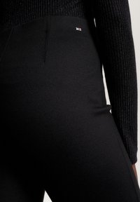 Juodos tamprės su elegantišku dizainu, su mažuoju prekės ženklu ant juosmens, pagamintos iš lygaus audinio su nepastebima siūle.