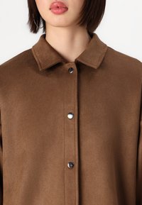 Camicia button-up in suede marrone con colletto, caratterizzata da bottoni a pressione argento e una finitura morbida e texturizzata. Design semplice e pulito.