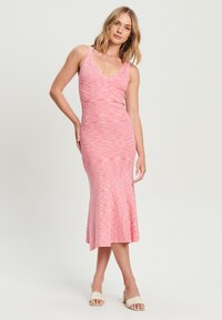 Ružová midi šaty s priliehavým telom a rozšíreným lemom, s výstrihom do V a rebrovanou textúrou. Nosené s svetlo béžovými sandálmi na otvorenú špičku.