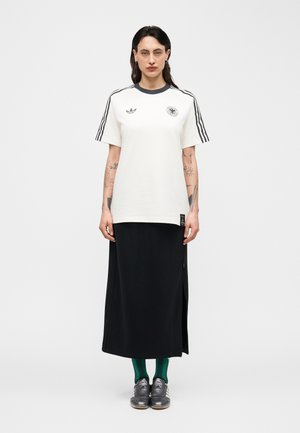 Person med langt mørkt hår iført hvit Adidas-T-skjorte med svarte striper og logoer, svart skjørt, grønne sokker og grå Adidas-sneakers, stående.