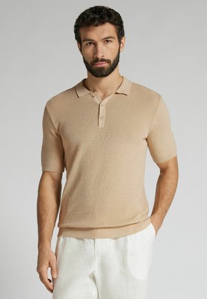 Mann mit Bart, der ein beiges kurzärmeliges Strick-Poloshirt und weiße Hose trägt, mit einer Hand in der Tasche vor einem schlichten Hintergrund stehend.