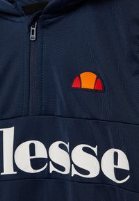 Marinefarbener Zip-Hoodie mit weißem "ellesse"-Logo, das eine kreisförmige, orange und rote gestickte Akzentuierung auf der Brust zeigt. Glattes, leichtes Material.