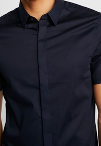 Námořnická modrá košile s krátkým rukávem vyrobená z hladkého materiálu, která má límec s knoflíky a jemně vyšitý logo na hrudi.