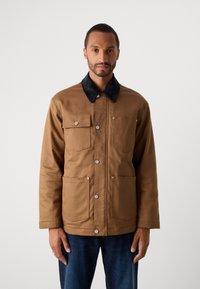 Hombre con una chaqueta marrón abotonada y un cuello negro, con cuatro bolsillos frontales, combinada con jeans azul oscuro, de pie frente a un fondo liso.