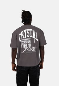 Šedé oversized tričko s krátkými rukávy. Na zádech se nachází výrazný bílý nápis "CRYSTAL" a grafika Arc de Triomphe a Eiffelovy věže.