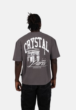 Šedé oversized tričko s krátkými rukávy. Na zádech se nachází výrazný bílý nápis "CRYSTAL" a grafika Arc de Triomphe a Eiffelovy věže.