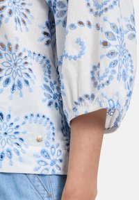 Witte blouse met blauwe bloemenborduring. Kenmerkt zich door gepofte mouwen en een geraffineerde manchette. Opmerkelijke details zijn onder andere open borduring en een decoratieve knoop.