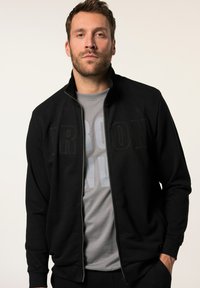 Chaqueta negra con cremallera y cuello alzado, con letras grandes de textura "IRON". Usada sobre una camiseta gris con letras en negrita.