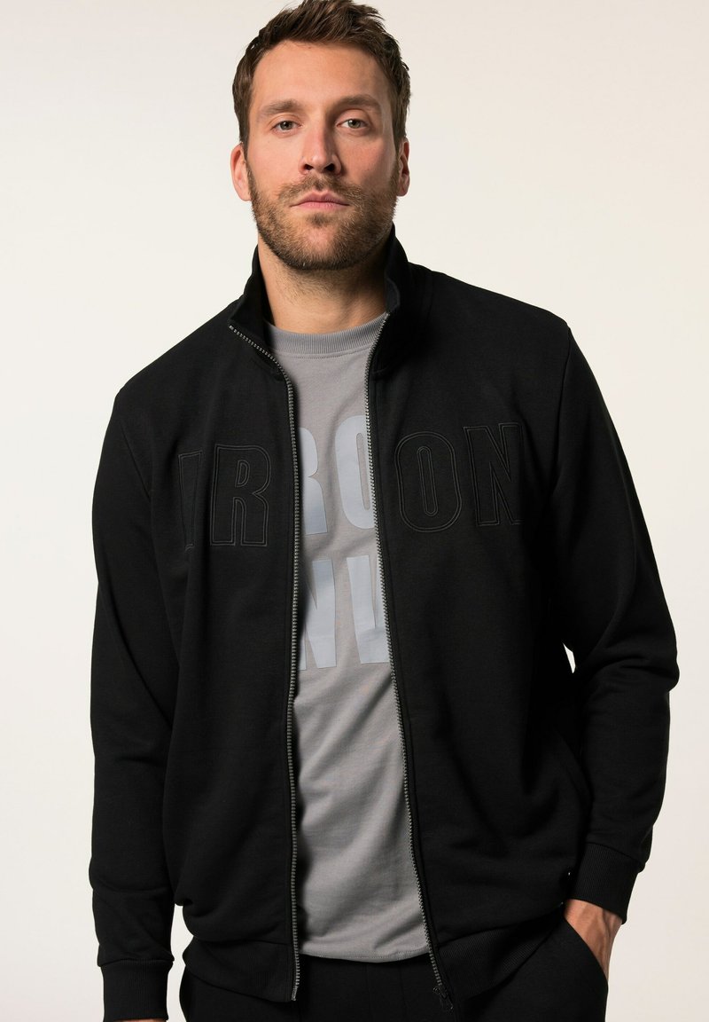Chaqueta negra con cremallera y cuello alzado, con letras grandes de textura "IRON". Usada sobre una camiseta gris con letras en negrita.