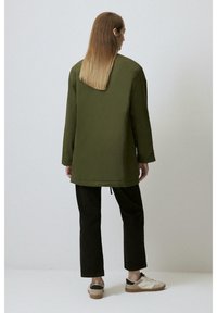 Jachetă verde cu mâneci lungi, cu o croială lejeră, cu fante laterale și o fustă rotunjită; asortată cu pantalonii negri și adidași în contrast.