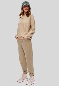 Beige trui met lange mouwen, beige relaxte broek en lichtgekleurde hoge sneakers. Een grijze pet maakt de outfit af.