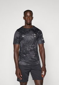 Un mannequin masculin portant un maillot de sport noir avec le logo de Tottenham Hotspur et un short noir, se tenant devant un fond blanc uni.