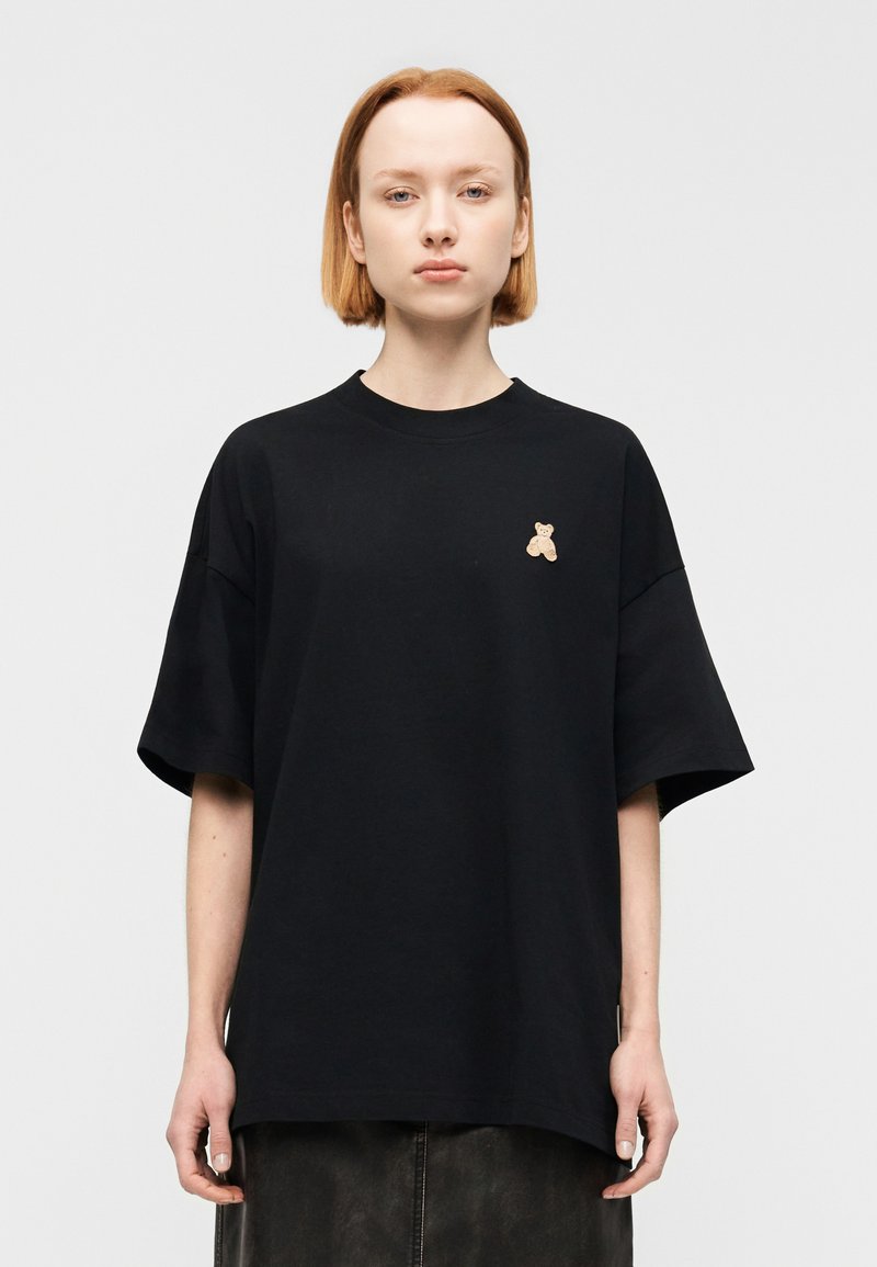 Μαύρο βαμβακερό t-shirt με χαλαρή γραμμή, στρογγυλή λαιμόκοψη και κοντά μανίκια. Διαθέτει μια μικρή κεντητή απεικόνιση αρκουδάκι σε μπεζ χρώμα στο στήθος.