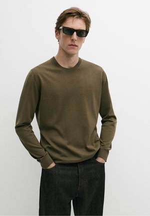Junger Mann mit schwarzen Sonnenbrillen, einem olivgrünen Langarmpullover und dunklen Hosen mit den Händen in den Taschen vor einem einfarbigen Hintergrund.