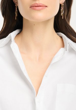 Frau trägt ein weißes Hemd mit offenem Kragen und langen, goldfarbenen Ohrhängern mit mehrfarbigen Steinen.