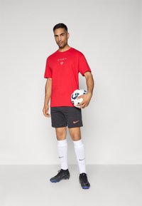 Rød atletik t-shirt med "ATLETI" logo, grå shorts med orange Nike swoosh, hvide sokker og sorte fodboldstøvler, mens han holder en mønstret fodbold.