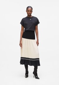 Black short-sleeve top with logo, paired with a pleated beige and black skirt and black ankle boots with a glossy finish.