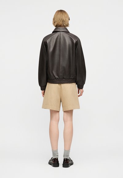 Άτομο με κοντά ξανθά μαλλιά που φοράει σκούρο καφέ δερμάτινο μπουφάν, μπεζ σορτς, γκρι κάλτσες και σκούρα καφέ παπούτσια, στεκεται με την πλάτη προς το μέρος μας.