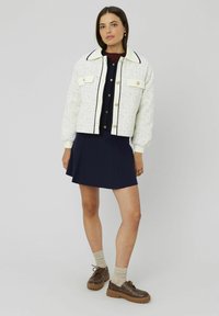 Veste blanche texturée avec des accents noirs, associée à une robe marine. Chaussures marron et chaussettes beiges complètent la tenue, mettant en avant les couches et le contraste.