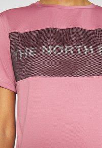 Camiseta deportiva de manga corta en color rosa con un panel de malla negra con el texto "THE NORTH FACE" en la parte frontal. Textura suave, ajuste relajado.