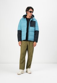 Uomo che indossa un giubbotto imbottito blu e nero, una maglietta nera, pantaloni verde oliva, sneakers bianche e occhiali da sole neri, in piedi contro uno sfondo bianco.