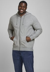 Haut à capuche gris zippé avec cordons, poches avant, poignets et ourlet côtelés. Porté sur un jean bleu, avec une texture lisse et semblable au coton.