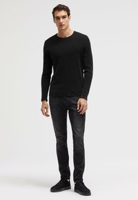 Black textured long-sleeve shirt paired with dark gray slim-fit jeans and black shoes, standing on a neutral background.