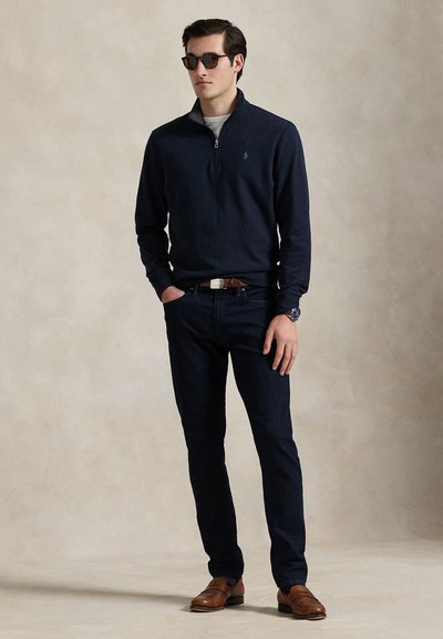 Homme portant un pull noir à demi-fermeture éclair, jean foncé, ceinture marron, chaussures marron, montre et lunettes de soleil, debout devant un fond clair uni.