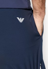 Temno modri atletski šortsi iz lahkega materiala. Imajo bel logotip na stegnu, stranske žepe in dihljive mrežaste podrobnosti.