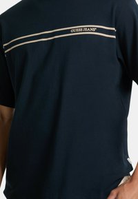 T-shirt in cotone blu navy con scollatura tonda. Presenta due strisce orizzontali cremmate e il logo "GUESS JEANS" al centro. Maniche corte.