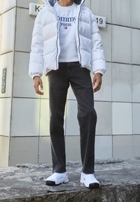 Chaqueta blanca acolchada, sudadera gris con el logo "Tommy Jeans", jeans gris oscuro, zapatillas de eslip blancas, de pie sobre una superficie rocosa.