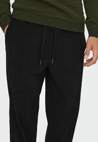 Schwarze Cordhosen mit glatter Textur, ausgestattet mit einem elastischen Taillenbund und Zugbändern. Die Hand des Models ist seitlich sichtbar.