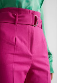 Fuchsia högmidjade byxor med bälte i midjan, veck och sidofickor; tyget ser slätt ut med en lätt lyster.