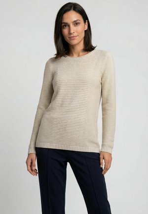 Vrouw met donker haar draagt een beige gestructureerde gebreide trui en een zwarte broek, staand tegen een effen lichtgrijze achtergrond.