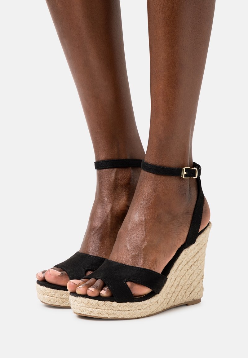 Pieds portant des sandales compensées en daim noir avec des brides à la cheville et des talons plateforme en jute tressé sur un fond clair uni.