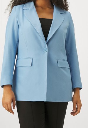 Mujer con blazer azul claro de un solo botón sobre una blusa negra y pantalones negros, de pie frente a un fondo liso.