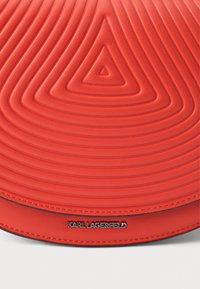 Punainen nahkainen pyöreä laukku, jossa on kohotettuja ympyräkuvioita, musta korostusnauha ja hopeinen logolevy, joka on painettu sivulle.